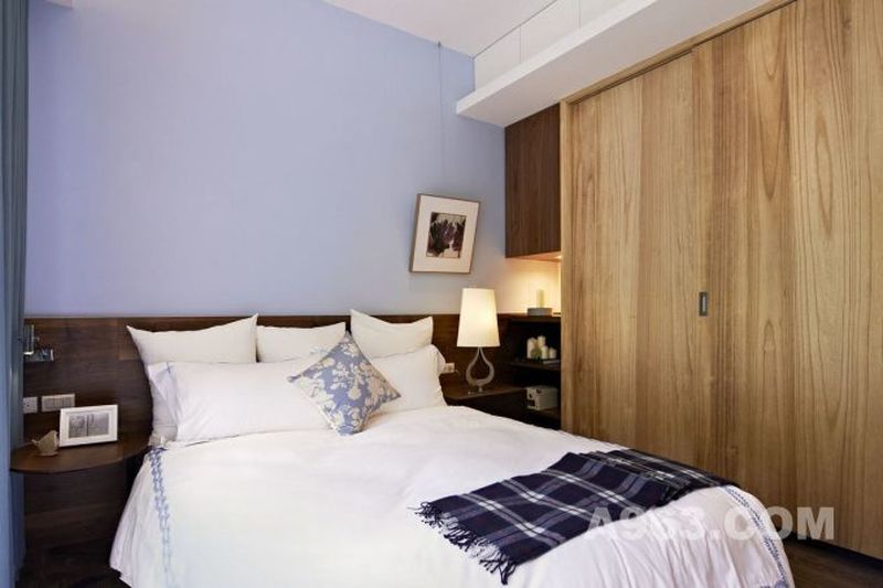卧室中放置着简约的大床，搭配上白色加淡蓝镶边的布面床品，贞洁而雅致。床头的背景墙则是以淡蓝色乳胶漆刷至而成，旁边则是自然原木橱柜设计，自然实用。
