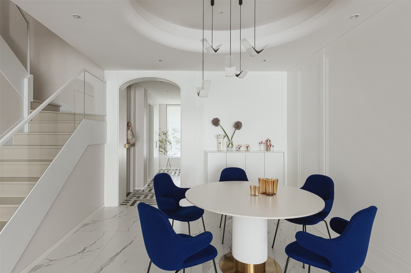 简约现代的客厅中，克莱因蓝色的餐椅在白色背景之下格外显眼，明丽优雅，柔和的弧度为用餐氛围更增美好的仪式感。餐厅中的圆形吊顶呼应了空间中的弧形元素，寓意一家人的团圆和美。