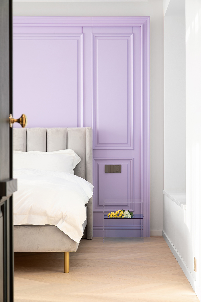 床头背景是一面香芋紫色轻法式护墙板，搭配渐变色极简亚克力床头柜和高级灰高背软包床，材质软硬结合，色调相辅相成，在悬浮吊顶和周边灯带的映照下，共塑一室温柔。